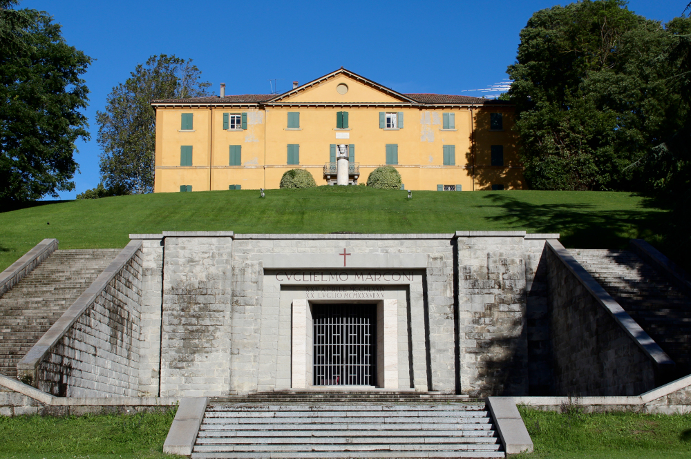 A three-storey yellow mansion stands on a hilltop. Built into the hillside below is the entrance to a grey stone mausoleum. The words “Guglielmo Marconi” are engraved over the door.