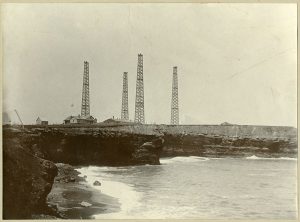 Four large aerial towers and a low station building atop cliffs at a seashore.