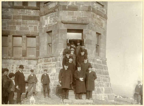 A group of men pose for a photo on the steps of a stone building. Young boys in caps, and one dog, watch the event.