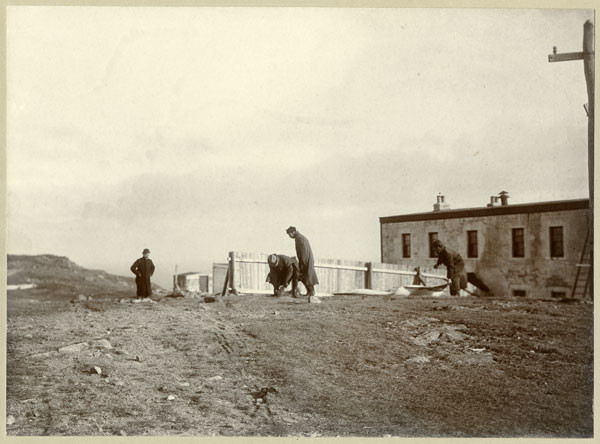 Four men set up a large kite on the ground. Behind them are a wooden fence and an old concrete building. In the distance are hilltops and the ocean.