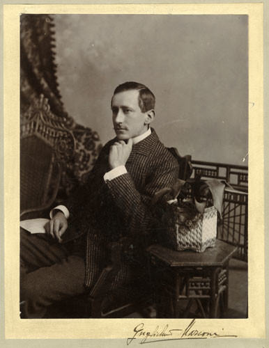 A young man with a moustache, wearing a suit jacket, sits posed for a formal portrait, his left hand beneath his chin.