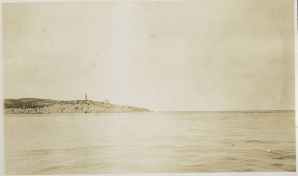 A calm ocean and big sky are separated by a low strip of land with a lighthouse tower.