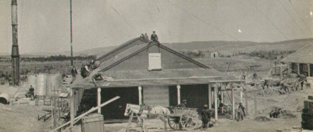 A single-storey wooden building with a horse and cart in front. Tradespeople work around the building and on the roof.