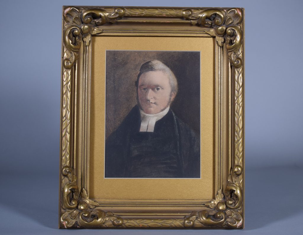 Portrait of William Abbott, charcoal, head and shoulders, adult. He has short hair and light brown sideburns, light grey eyes. He is wearing a black minister’s robe and a white collar. The background is dark grey-brown. The portrait is set in a gilded woodwork frame with gold mat.