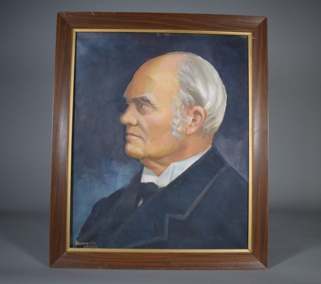 Profile portrait of an elderly John Joseph Caldwell Abbott, oil, colour, head and shoulders. His hair and sideburns are white and grey. He is wearing a white shirt with black tie and jacket. The background is dark blue-grey. The artist’s signature, “Jeannette Wales”, can be seen in the lower left corner. The portrait is set in a simple wood frame with a narrow gold mat.