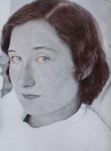 Black and white photograph with coloured details of an adult woman; head and shoulders. She is looking at the camera, in three-quarters profile. Her pale blue eyes are reddened, and her hair is reddish-brown. She has thin lips and eyebrows and freckles on her cheeks and nose. She is wearing a white sweater with a mid-neck collar.