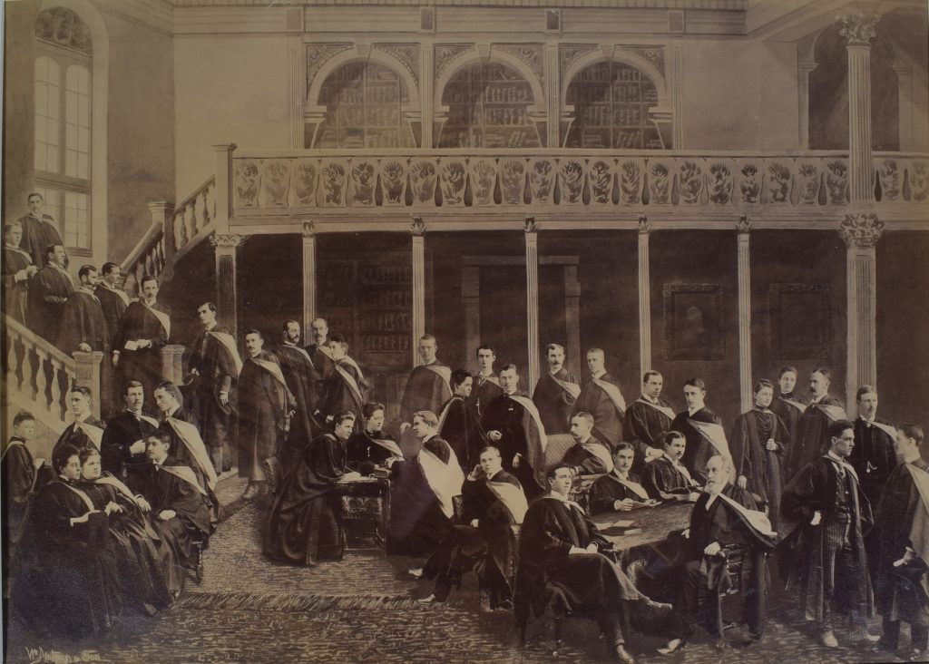 Photograph of a class of young adults, men and a few women, sepia. There are 35 students arranged around a room. They are all wearing black graduation gowns with a white sash with dark edges over the left shoulder. The men have short hair and the women have their hair tied behind their heads. They are in a room with a large carpet on the floor, in front of a balcony, stairs with columns and large bookshelves.