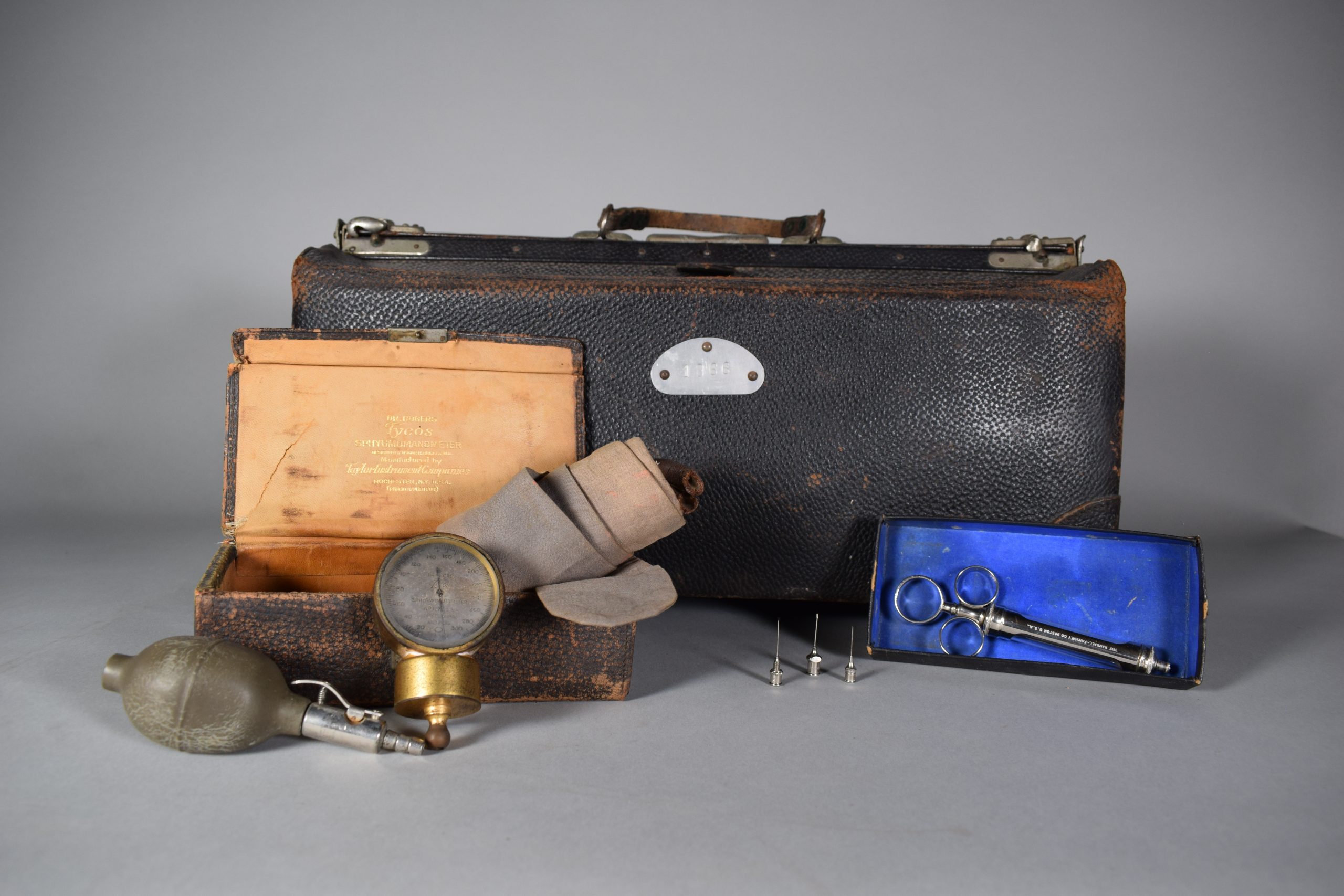 Colour photograph of a doctor’s bag and instruments. At the back there is a large rectangular black leather bag with a handle in the centre and clasps on both sides. The bag is slightly damaged at the corners. In front of the bag and to the left is a smaller case, also made of black leather. It is open, showing a beige interior. The case contains a large beige-grey cotton bandage. A blood pressure bulb and a gold-coloured sphygmomanometer lean against the case. To the right, there is an old syringe in a cardboard box and three needles can be seen to its left.