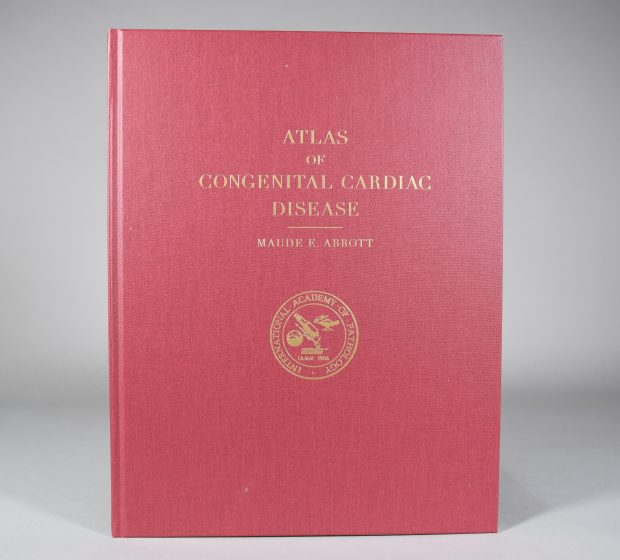 Colour photograph of Maude Abbott’s Atlas. It is red with the inscription “Atlas of Congenital Cardiac Disease - Maude E. Abbott” in gold lettering. Under the title is the gold seal of the International Academy of Pathology: the name of the academy encircles images of a globe, a microscope and the lamp of learning, above the inscription “IAMM 1906”.
