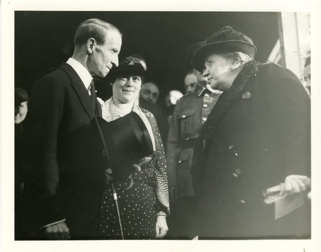 Photographie en noir et blanc de Maude Abbott et Lord Tweedsmuir lors de la cérémonie de commémoration de Sir John Joseph Caldwell Abbott en 1936. Lord Tweedsmuir, à gauche, est vu de profil. Il a les cheveux courts, séparés sur le côté et porte un veston noir. Il échange un regard avec Maude Abbott, à droite dans la photo. Elle est âgée et porte un manteau noir avec un col en fourrure. Derrière Lord Tweedsmuir, une femme avec une robe foncée à pois blancs et un chapeau chic le regarde attentivement. On aperçoit la foule derrière eux.