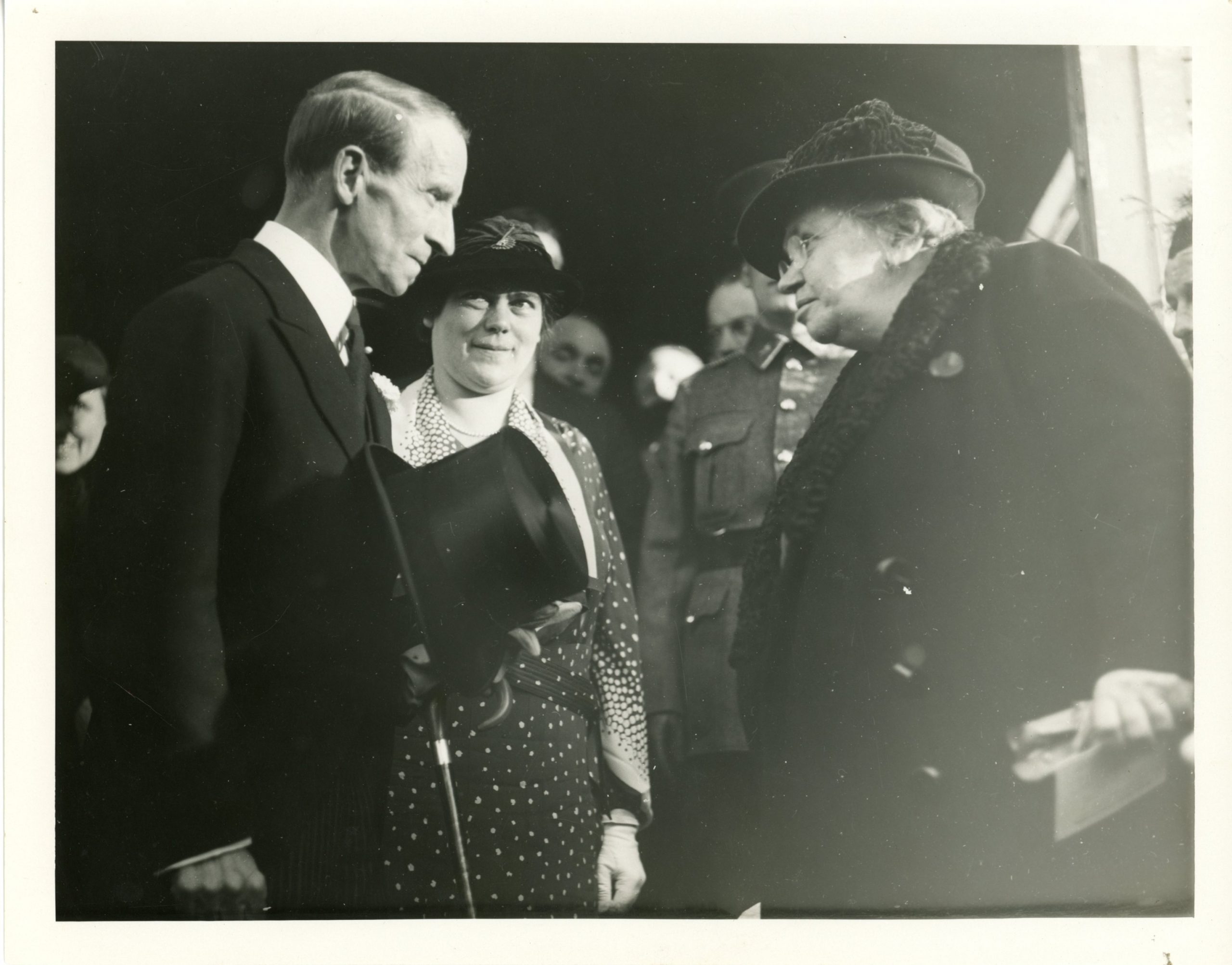 Black and white photograph of Maude Abbott and Lord Tweedsmuir during the Sir John Joseph Caldwell Abbott memorial dedication ceremony in 1936. Lord Tweedsmuir, on the left, is seen in profile. He has short hair, parted on the side, and is wearing a black jacket. He is exchanging glances with Maude Abbott, on the right in the photo. She is elderly and is wearing a black coat with a fur collar. Behind Lord Tweedsmuir, a woman in a dark dress with white polka dots and a stylish hat is looking at him attentively. The crowd can be seen behind them.