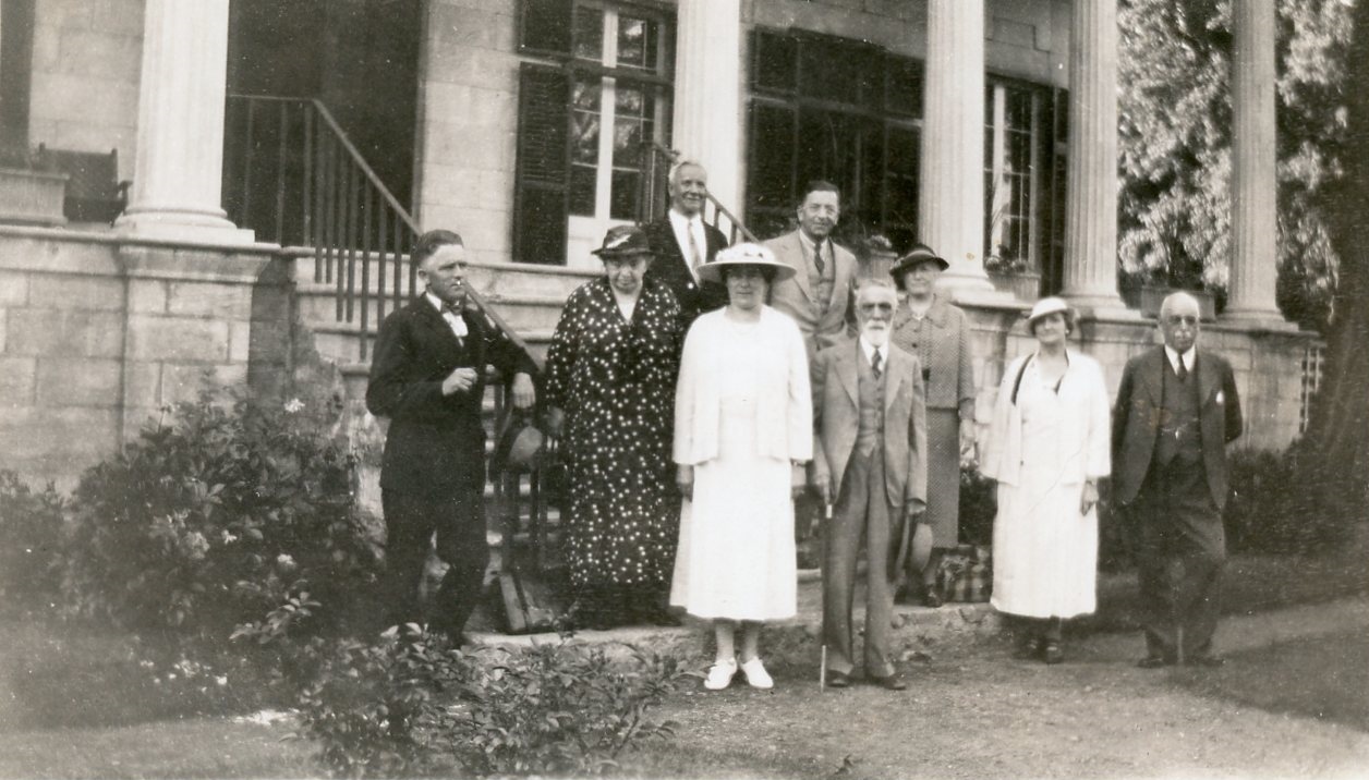 Black and white photograph of the nine founding members of the Historical Society of Argenteuil County. They are standing in front of and on the stairs of a large stone house with columns and dark shutters. Maude Abbott is second from left, wearing a black dress with white polka dots and a dark hat. The other three women are wearing smart dresses and hats and the five men are wearing suits.