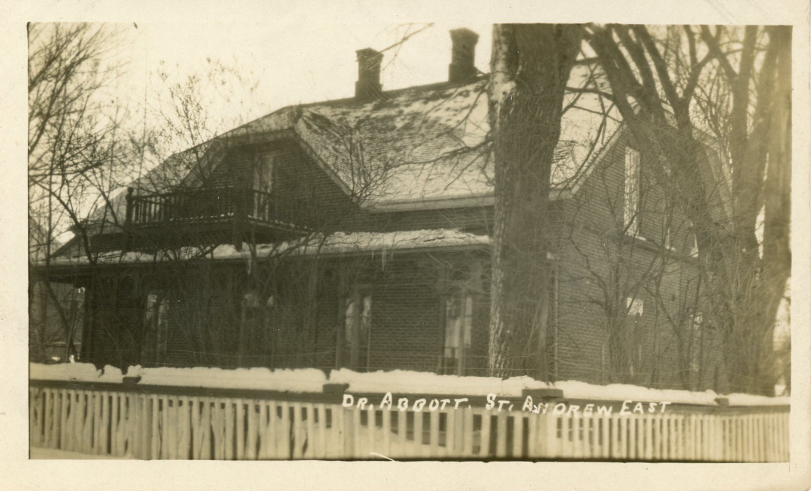 Photograph of Elmbank, Maude Abbott’s home, in winter, sepia. It is a two-storey brick house with a gable roof, two chimneys and a gallery on each floor. There is a white wooden fence in front of the house, which is surrounded by leafless trees.