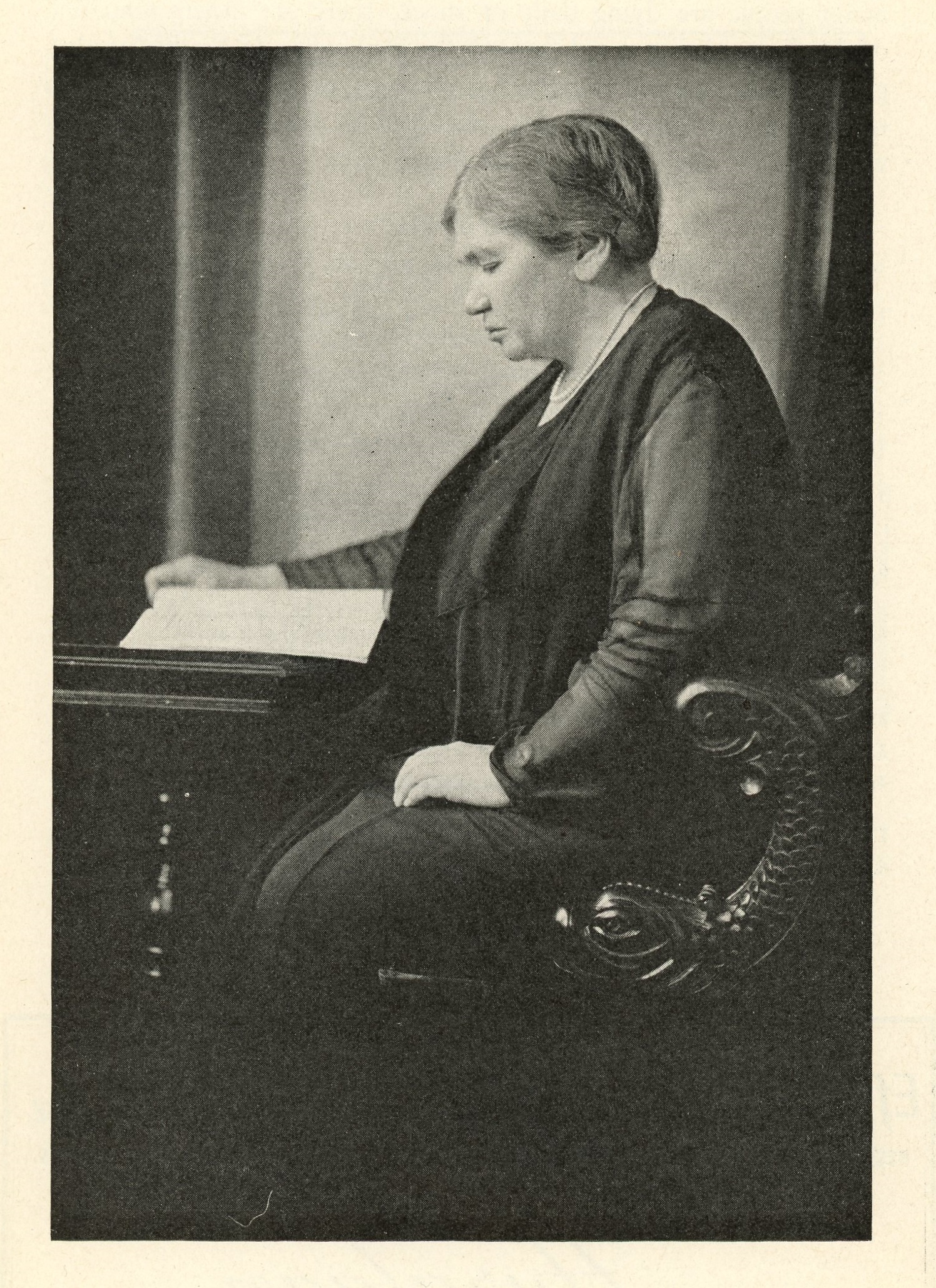 Photograph of an elderly Maude Abbott, black and white. She is seen in profile, looking concentrated, reading a book at a wooden desk and seated on a carved wooden chair. She is wearing dark clothes and a pearl necklace, and her hair is tied behind her head.