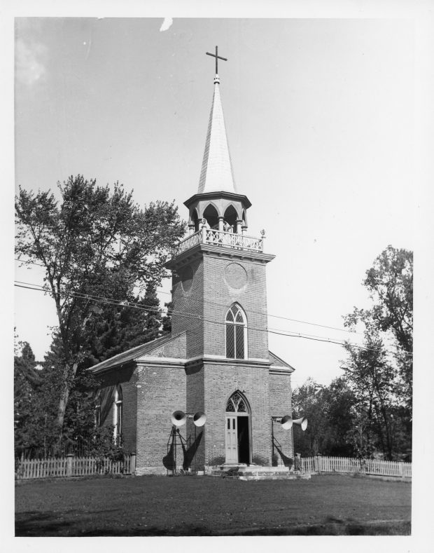 Black and white photo of Christ Church. The church is made of brick, the double door in the centre is open on one side and topped by a lancet window. Another window of the same shape can be found in the bell tower. The openings in the bell tower are lancet-shaped and it is topped by a tall spire crowned with a black cross. Two loudspeakers are mounted on either side of the door and there are many trees behind the church.