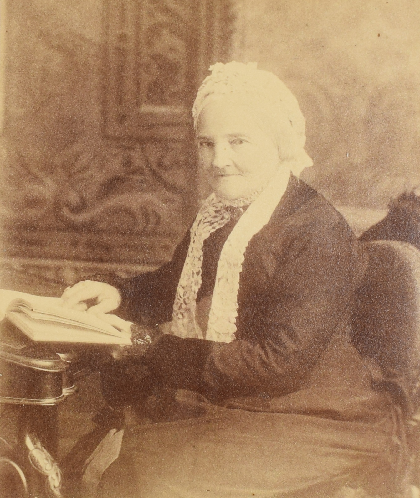 Sepia photograph of an elderly Frances Mary Smith, 1880, sitting, from the knees up. She is facing slightly left and looking at the camera with a calm expression. Her left hand rests on an open book on a desk. She is wearing a black dress and a white lace cap. Her white hair is tied behind her head, under the cap.