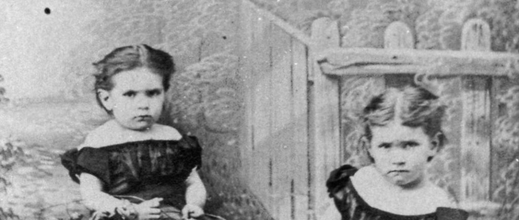 Black and white photograph of Maude and her sister Alice as children. They are sitting by a fence and gazing seriously at the camera. They are wearing dresses with bare shoulders and dark puffy sleeves. Maude, on the left, has dark hair tied behind her head and wears a flower on her right arm. Alice, on the right, has light hair, also tied behind her head. They are sitting behind an arrangement of flowers, two hats and three rocks on the lawn.