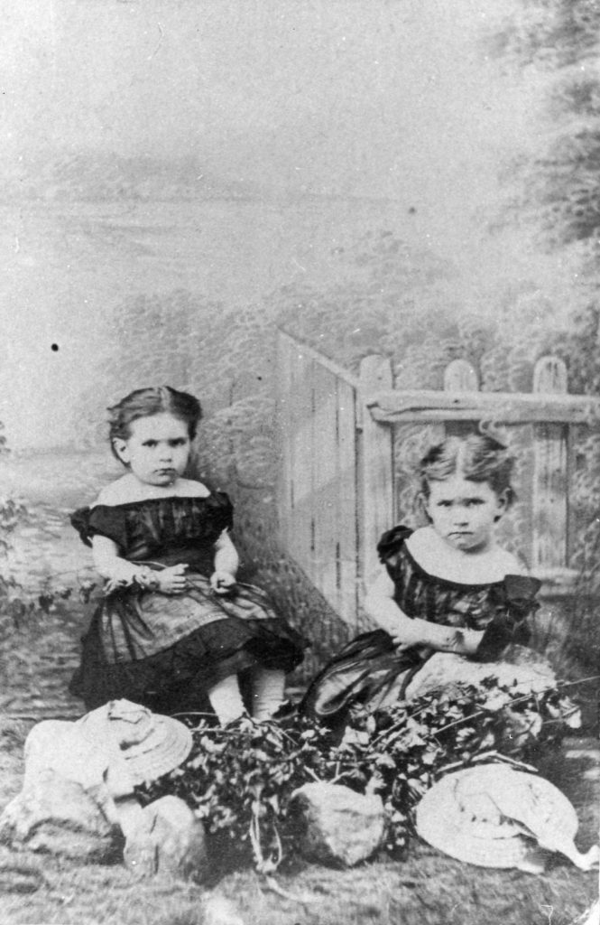 Black and white photograph of Maude and her sister Alice as children. They are sitting by a fence and gazing seriously at the camera. They are wearing dresses with bare shoulders and dark puffy sleeves. Maude, on the left, has dark hair tied behind her head and wears a flower on her right arm. Alice, on the right, has light hair, also tied behind her head. They are sitting behind an arrangement of flowers, two hats and three rocks on the lawn.