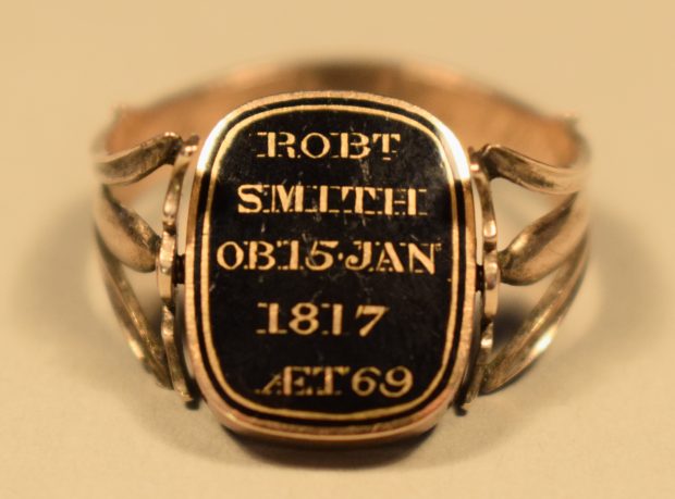 Colour photograph of the face of a rose gold ring. The ring is gold coloured and its face is decorated with gold lettering and border on a black background, bearing the inscription “ROBT SMITH OB 15 JAN 1817 AET 69”. The background of the photo is beige.