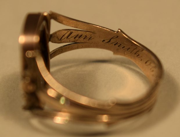 Colour photograph of a rose gold ring, inside left side. The ring is gold coloured and “Ann Smith” can be seen engraved on the shank (see photograph 4.1.3 for the continuation). The background of the photo is beige.