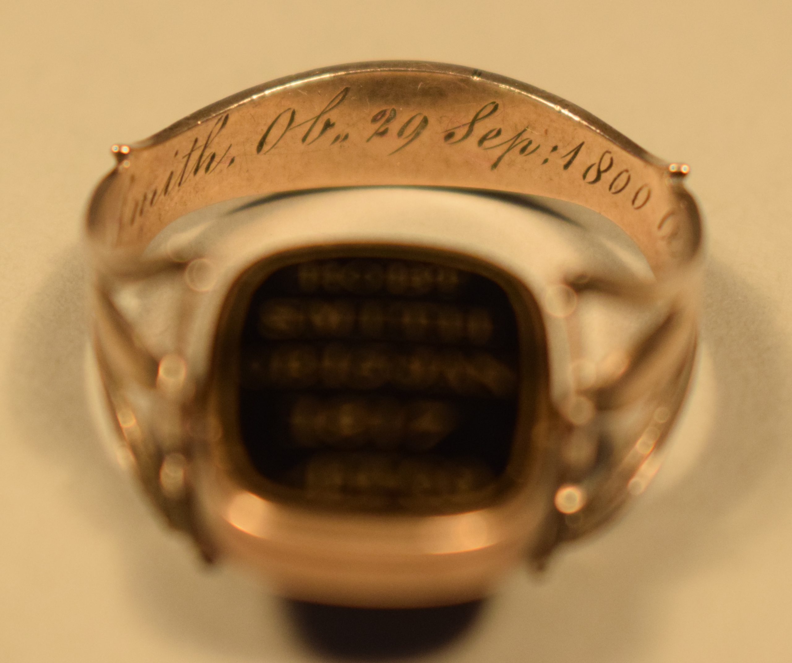 Colour photograph of a rose gold ring, inside back. The ring is gold coloured and “Smith Ob,, 29 sep : 1800” can be seen engraved on the shank (see photograph 4.1.4 for the continuation). The background of the photo is beige.