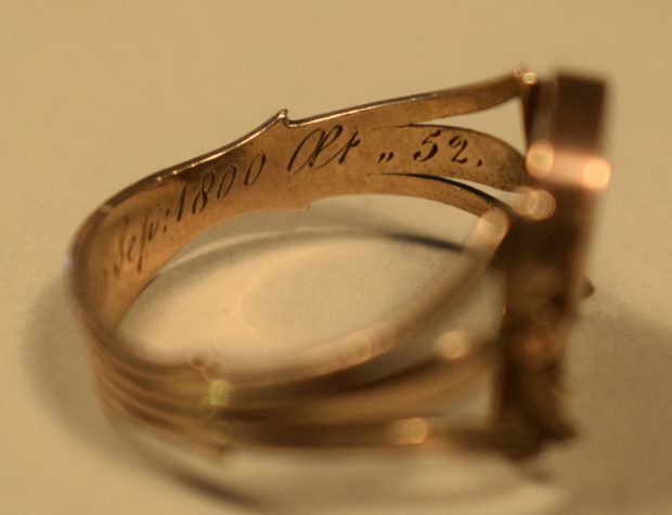 Colour photograph of a rose gold ring, inside right side The ring is gold coloured and “Sep : 1800 aet ,, 52” can be seen engraved on the shank. The background of the photo is beige.