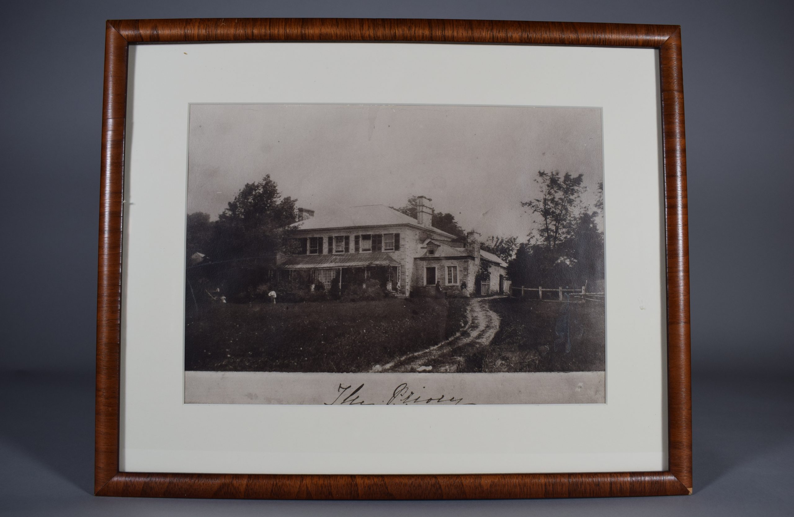 Black and white photograph of a two-storey house surrounded by trees in summer. It is made of grey brick with a large gallery. The upper storey has five windows with dark shutters and there are two chimneys on the roof. The side section of the house is slightly set back and has only one storey. Two women are standing in front of the side section, a man is sitting on the stairs and another is in the yard in front of the house. Below the photo, “The Priory” is written in black ink. The photograph is displayed in a simple narrow wooden frame with a white mat.