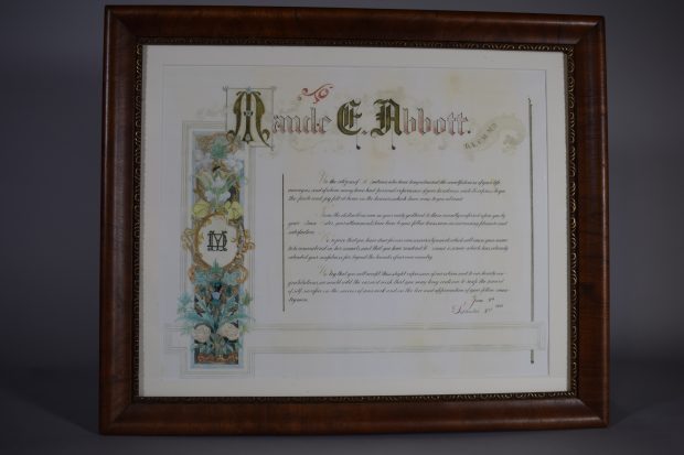 Certificate of recognition; colour. At the top, “Maude E. Abbott” with raised initials and a scroll to the right with the letters “B.A., C.M., M.D.” On the left, a banner with flowers in a black frame surrounds a golden circle with the overlapping letters “MD”.