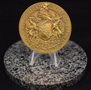 Colour photograph of the Lord Stanley Gold Medal awarded to Maude Abbott in 1888, reverse face. The medal is gold in colour and shows a coat of arms, surrounded by the inscription “Presented by His Excellence The Governor General”.