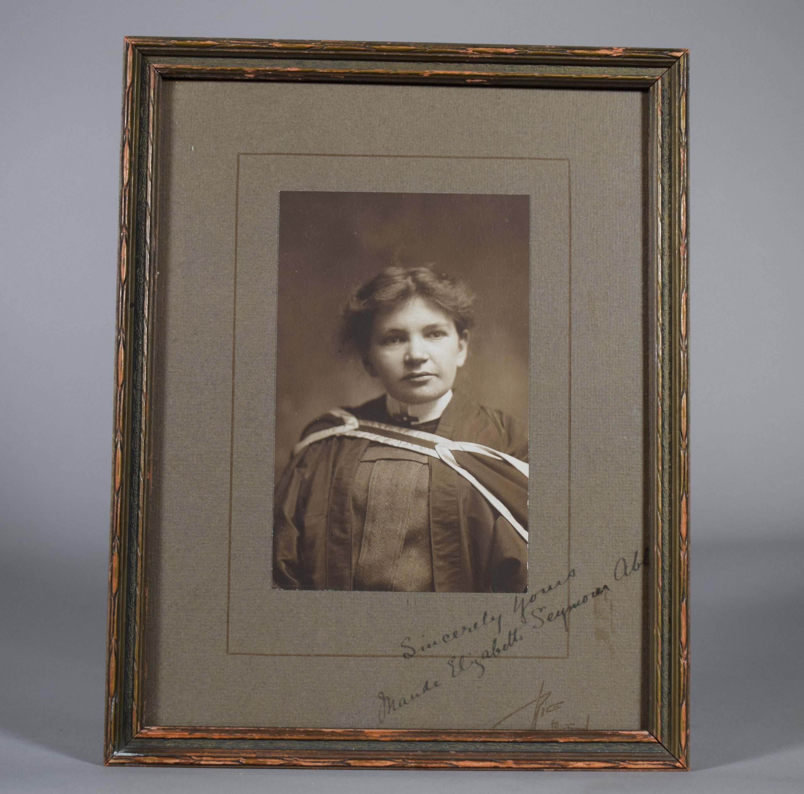 Black and white photograph of Maude Abbott as a young adult, from the waist up. She is wearing a graduation gown. Her dark hair is tied behind her head and she is looking slightly to her left. There is a signature in the lower right corner, in ink: “Sincerely Yours. Maude Elizabeth Seymour Abbott”. The printer’s signature appears below this: “Rice. Montreal”.