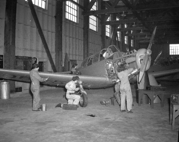 Black and white photograph showing five women mechanics in a hangar, where they are working on an airplane. A sixth person is sitting in the cockpit.
