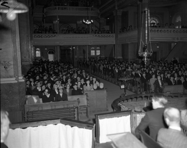 Black and white photograph showing members of the Cercle des fermières. In the main section of the auditorium, women are sitting on benches, on either side of the aisle. In front of them, three men appear to be making a presentation. More women are watching the proceedings from seats in the balcony.