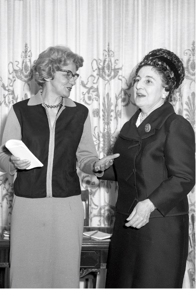 Black and white photograph of Adrienne Choquette and Claire Roy in conversation. The two women are standing in front of a desk
