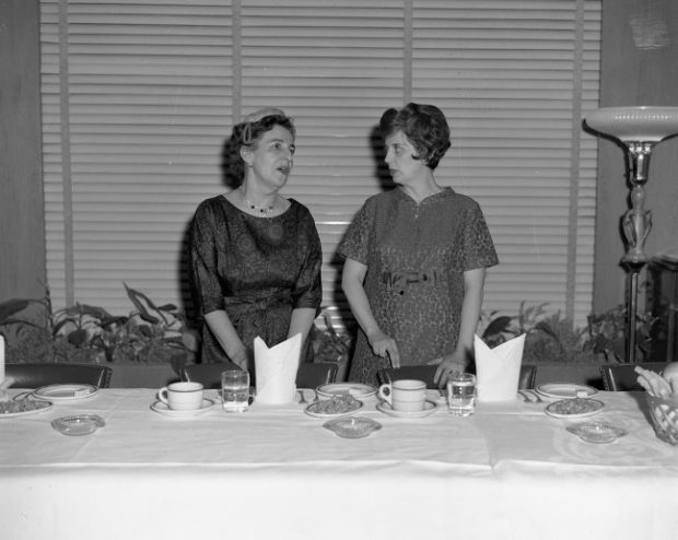 Black and white photograph of two women standing behind a table. Claire Roy (left) is talking to the other woman. There are dishes on the table in front of them, and a lamp can be seen on the right.