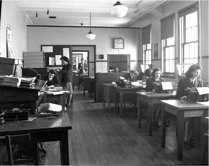 Black and white photograph showing women contributing to the war effort. On the right, several women are sitting at tables where they are typing letters. On the left, one woman is sitting at a desk covered with papers and another is looking through a filing cabinet. At the back, a man standing at a desk can be seen through the open door of an adjoining room.
