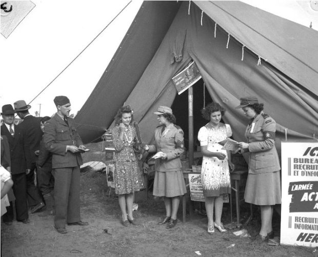 Black and white photograph showing four women, two of whom are in uniform, talking in front of a tent at an army recruitment centre. On the left, a man in uniform is standing in front of three other people.