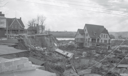 Black and white photograph of the aftermath of a landslide. Surviving houses are visible on the left of the photograph. On the right, a house impacted by the disaster lies among the ruins.