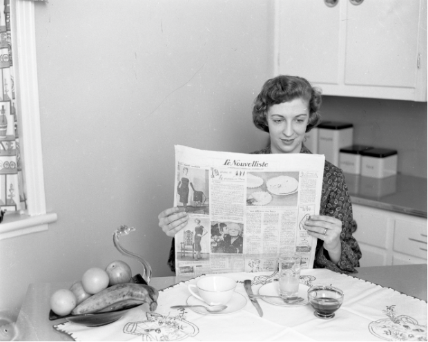 Black and white photograph of a woman sitting at the kitchen table, reading Le Nouvelliste. A cup, two glasses, two spoons, a knife and a swan-shaped bowl with fruit are on the table in front of her.