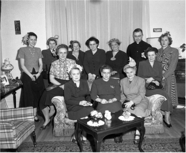 Photograph showing members of the Mauricie branch of the Ligue catholique féminine, along with a member of the clergy. Three women are seated on a sofa, with eight other women and the clergyman behind them. There is a coffee table in front of them and a chair to their right.