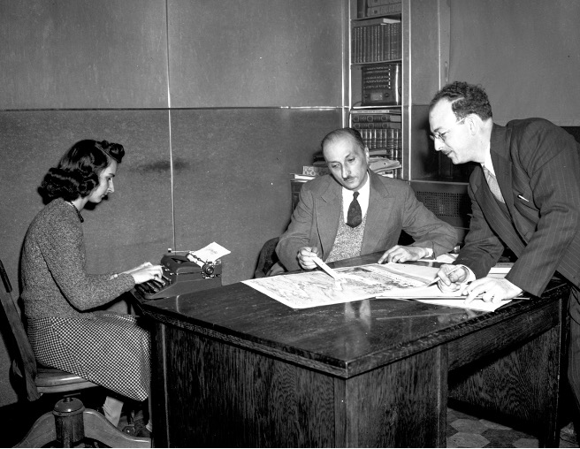 Black and white photograph of a woman transcribing a conversation between Managing Director Émile Jean and Local Advertising Manager Jos Lefebvre. Seated at a desk, Jean points to a map while Lefebvre, standing to the right of the desk, looks on.