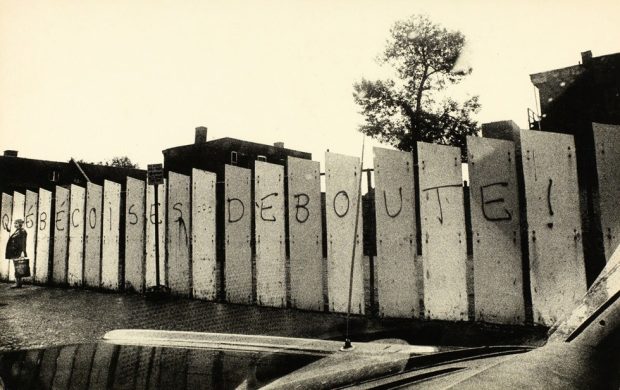 Black and white photograph showing a row of fence panels painted with the words “Québécoises Deboute!” A woman is standing next to the fence.