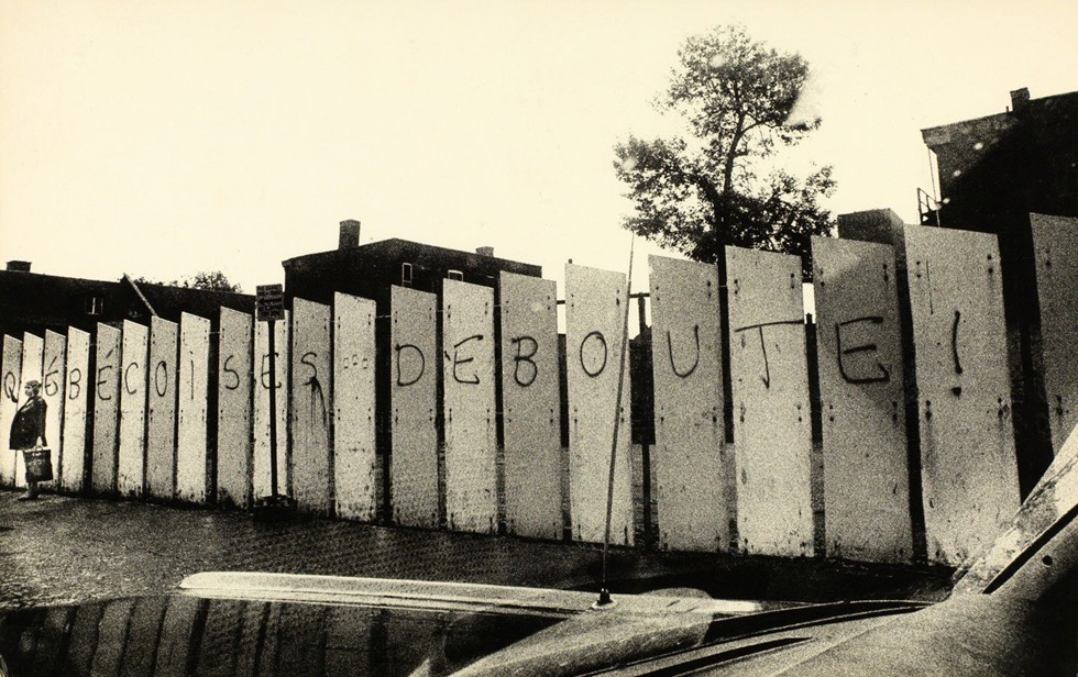 Black and white photograph showing a row of fence panels painted with the words “Québécoises Deboute!” A woman is standing next to the fence.