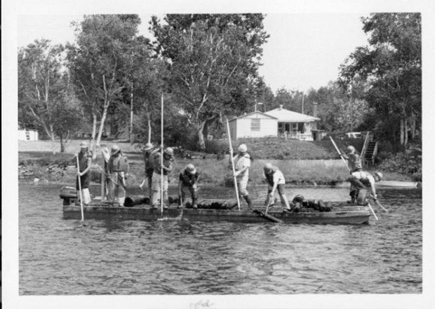 Black and white photograph showing people working on a log drive on the Saint-Maurice River in 1987. Nine people are standing on a pontoon, holding poles.