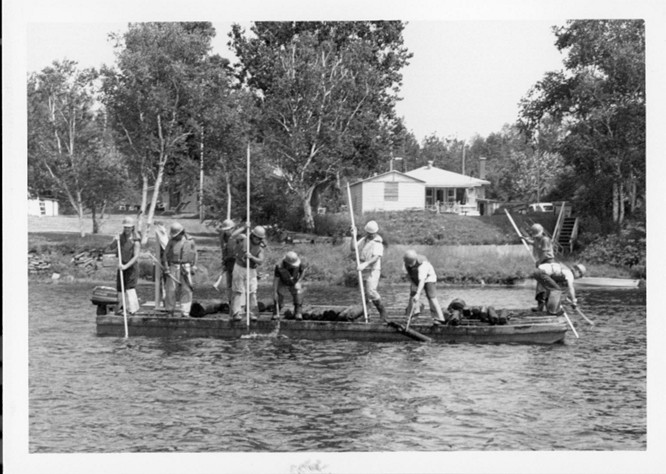 Black and white photograph showing people working on a log drive on the Saint-Maurice River in 1987. Nine people are standing on a pontoon, holding poles.