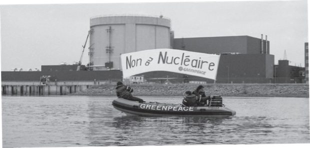 Black and white photograph of three Greenpeace activists in an inflatable boat on the St. Lawrence River, in front of the Gentilly-2 Nuclear Generating Station. They are holding up a banner with the words “No to Nuclear.”