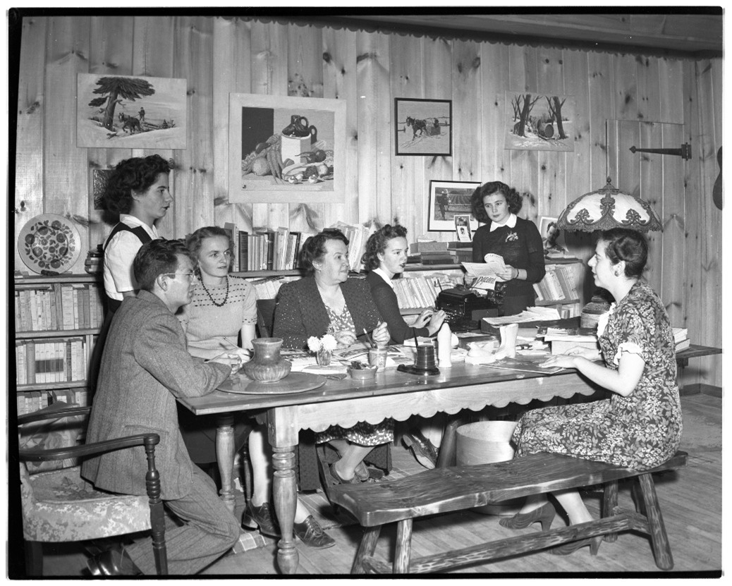 Black and white photograph of six women and one man seated around a table inside a home in 1946.