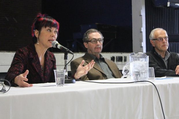 Colour photograph of a woman and two men sitting at a conference table. Sitting on the left, the woman is dressed in red and black. She is speaking into a microphone. In addition to the microphone, a drinking glass and a water pitcher are visible on the table.