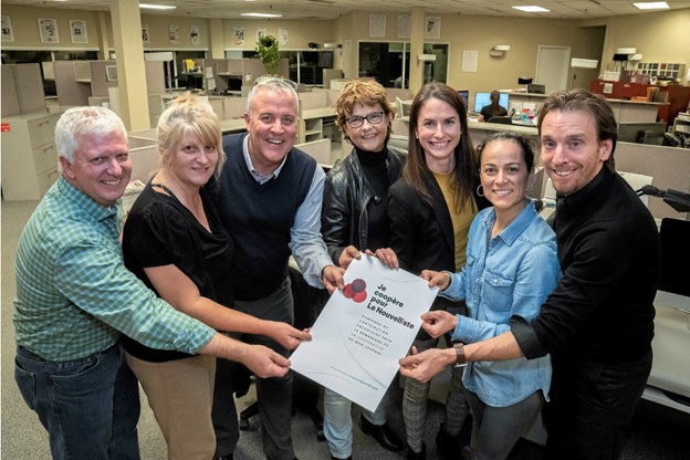 Colour photograph of seven people, three men and four women, standing in the newsroom. They are holding a sheet of paper that reads: “I cooperate for Le Nouvelliste.”