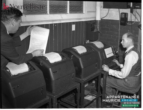 Black and white photograph showing four industrial typewriters and two men in a newsroom. One man is standing on the left, reviewing a text that has just been printed out. The other man is sitting on the right, typing on one of the machines. Electronic devices are mounted on the wall at the back of the room.
