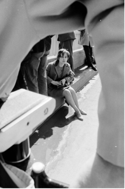 Black and white photograph of Marina Roy sitting on a sidewalk, with men standing on either side of her.